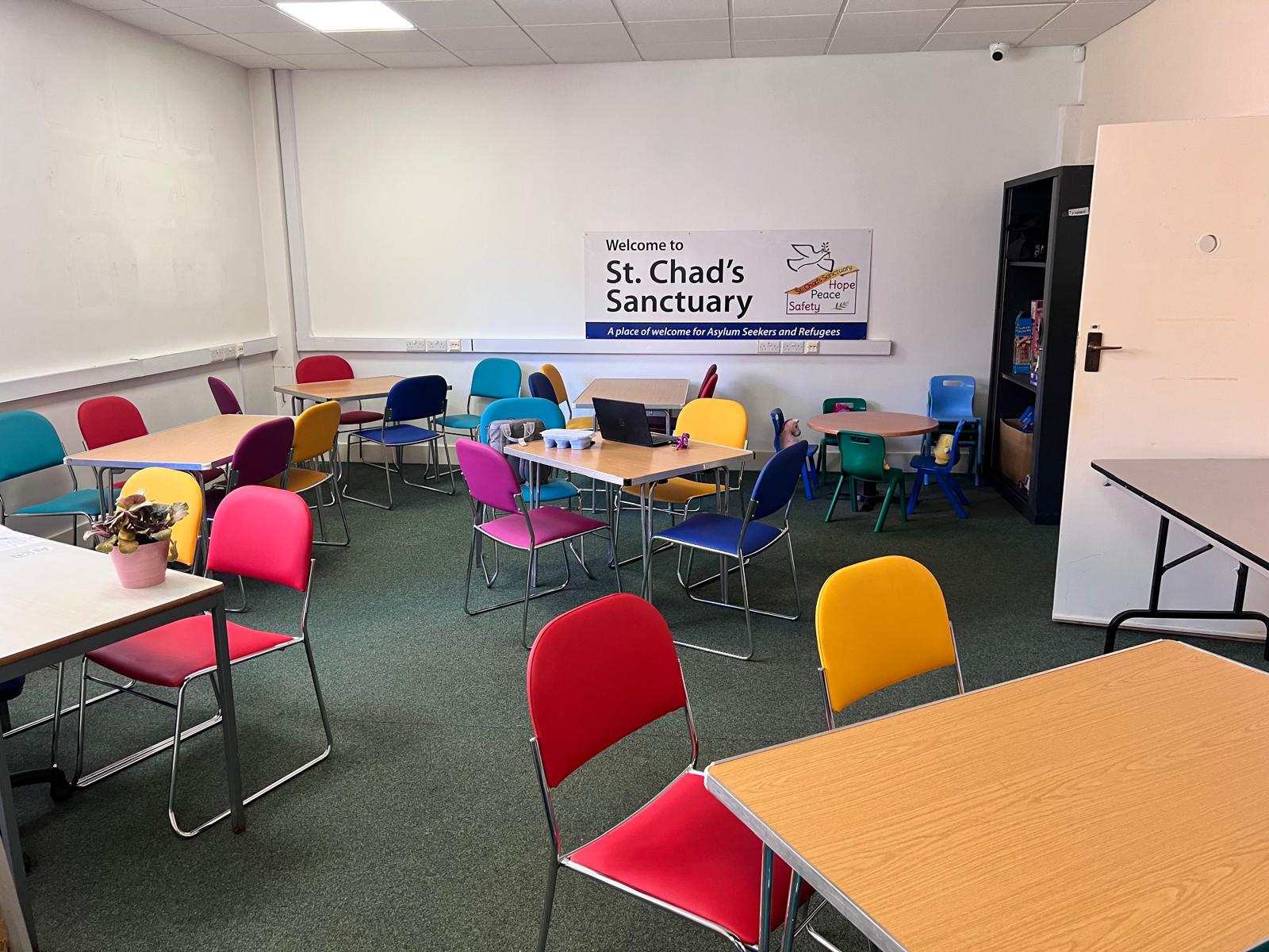 The drop-in room at St Chad's Sanctuary: tables and brightly coloured chairs under a 'Welcome to St. Chad's Sanctuary' sign.