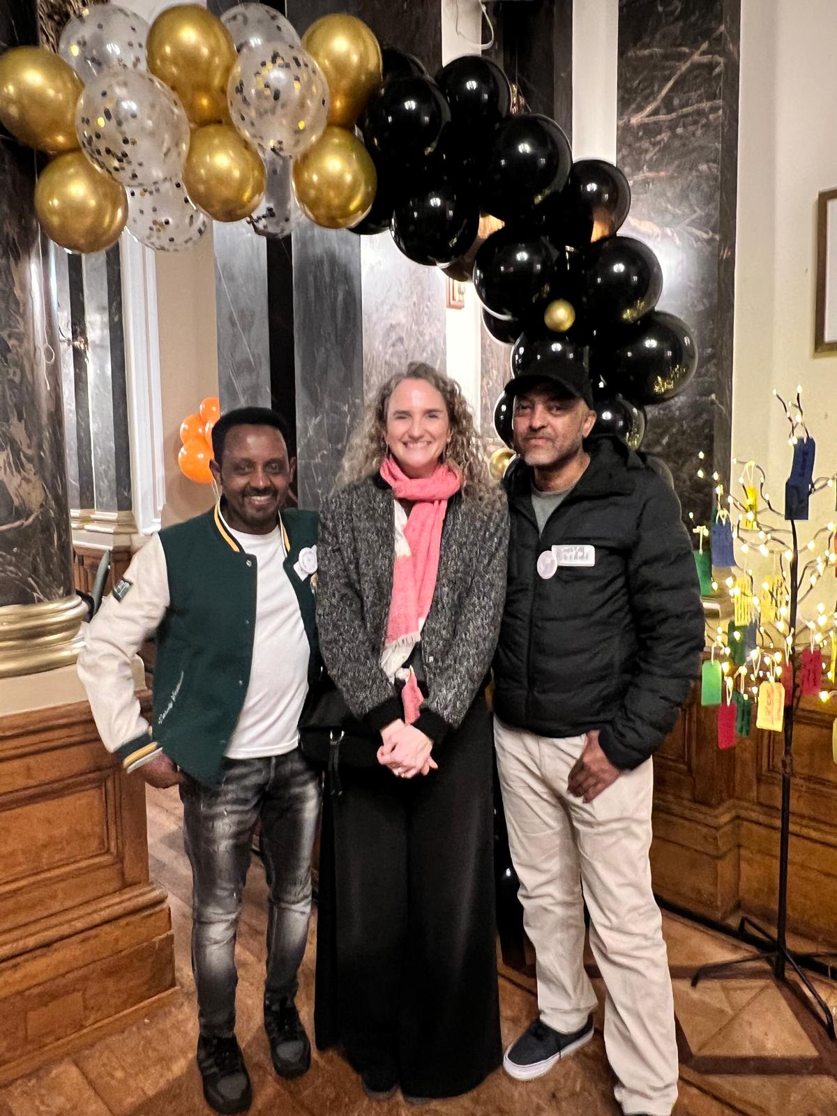 Three people smiling together at an event, with gold and black balloons behind them.