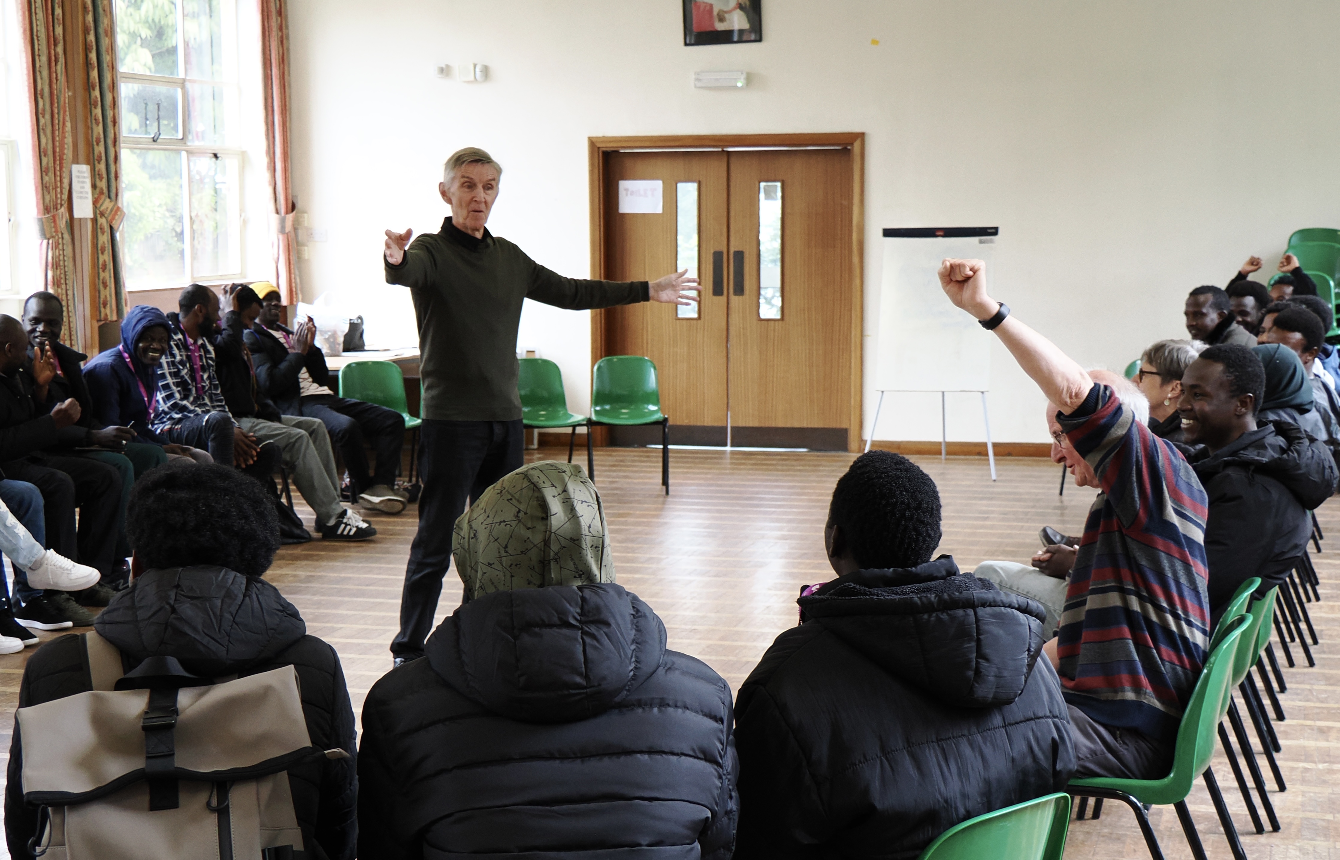A volunteer leads a Fun with English session in a hall, with people seated in a circle joining in.