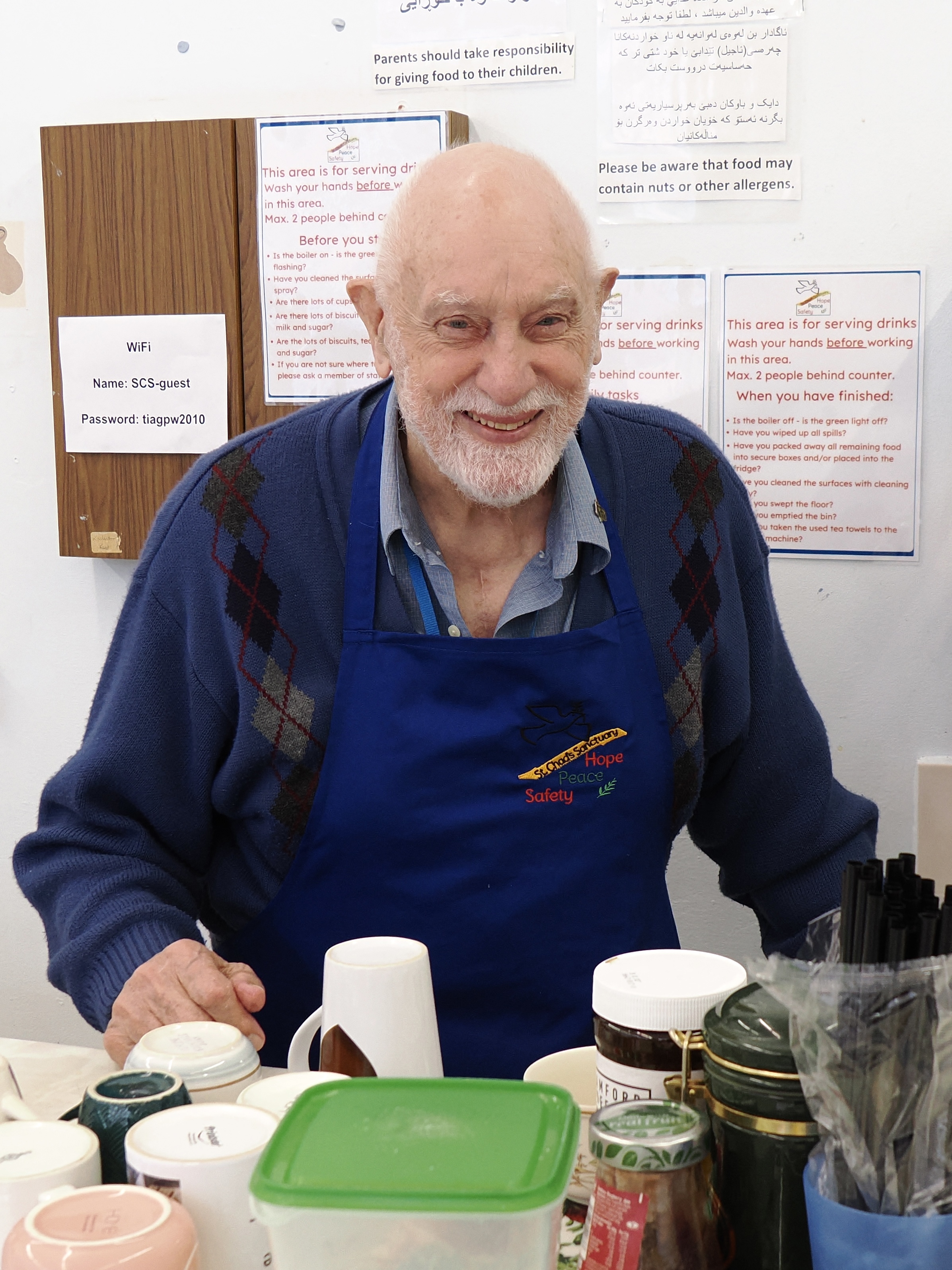 A smiling volunteer in a blue apron at the tea and coffee counter.